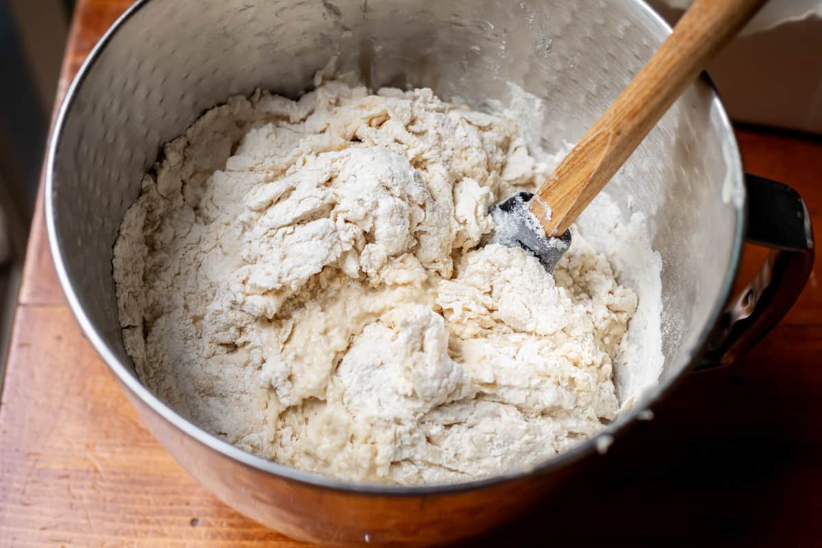 a metal mixing bowl with a spoon stirring in the flour into the mixture before kneading.
