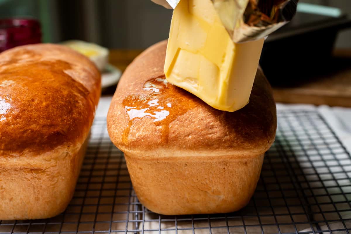 a whole stick of butter being rubbed on the top of a freshly baked loaf of homemade bread.