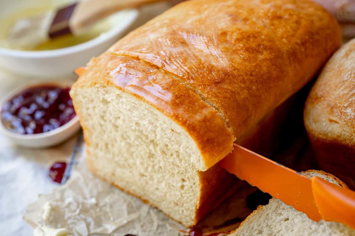 a bread knife carefully cutting a thick slice from a whole loaf of homemade white bread.