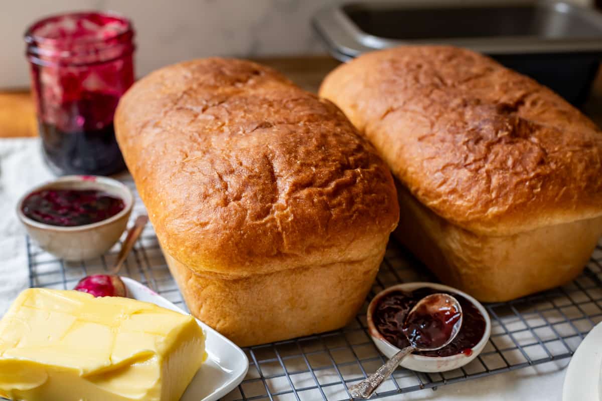 two loaves out of the pan of uncut fast white bread with dishes of butter and jam next to them.