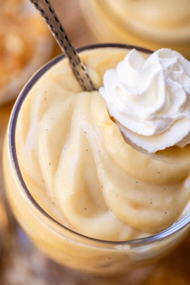 single glass cup of creamy vanilla pudding with a spoon and a dollop of whipped cream.