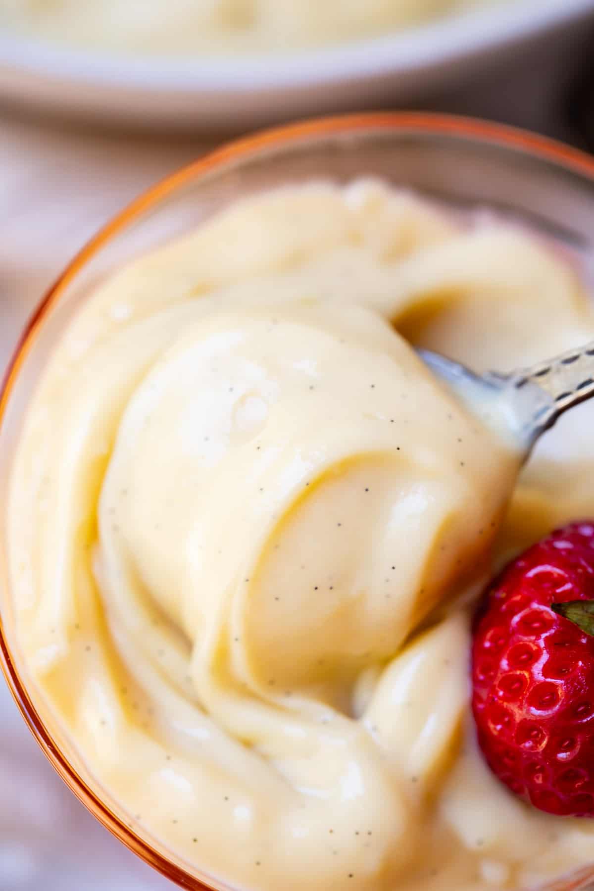 Close up of a spoon lifting a bite of vanilla pudding from a pink glass serving dish.