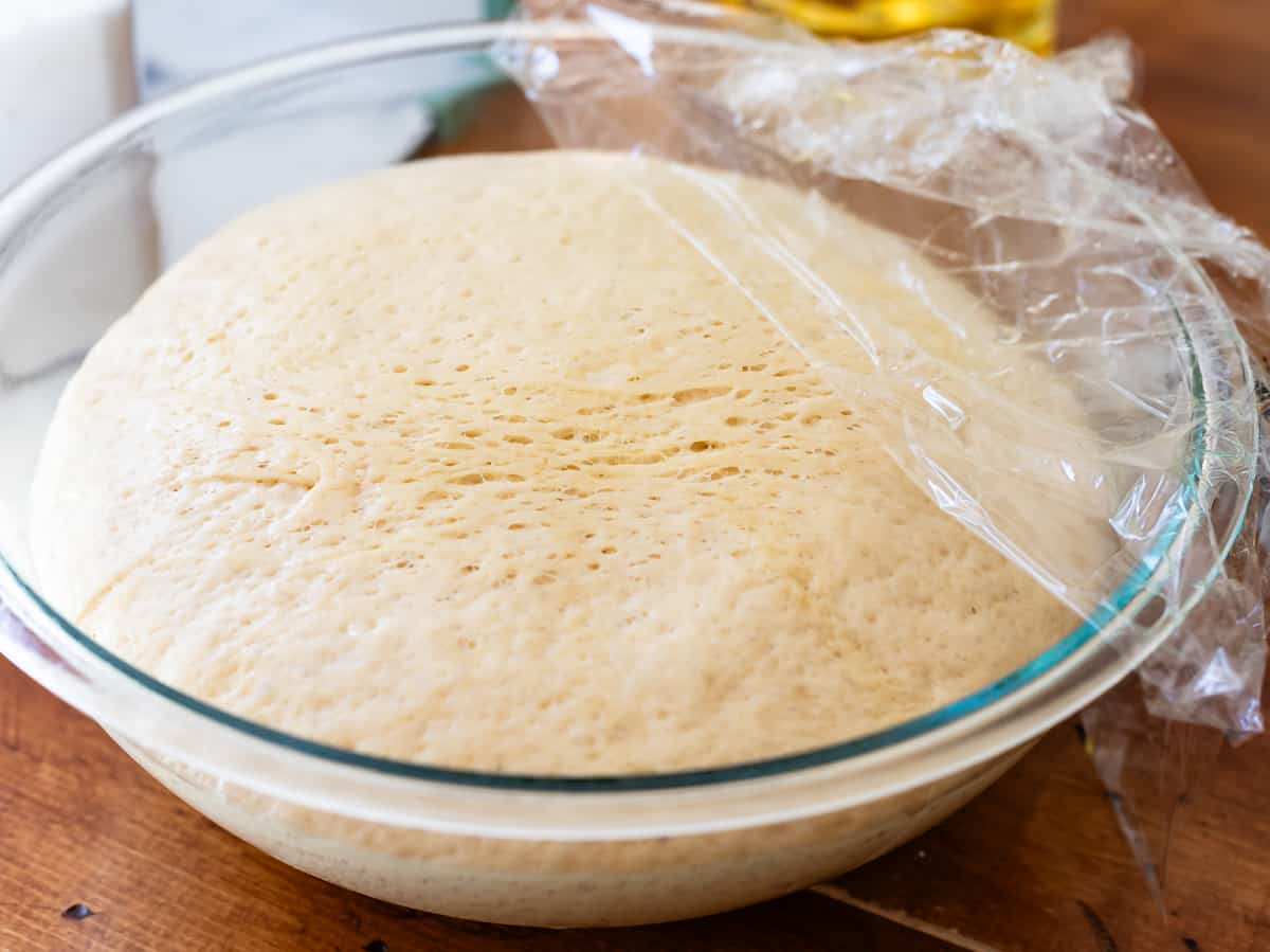 risen homemade donut dough in a glass mixing bowl with plastic wrap pulled off the top.