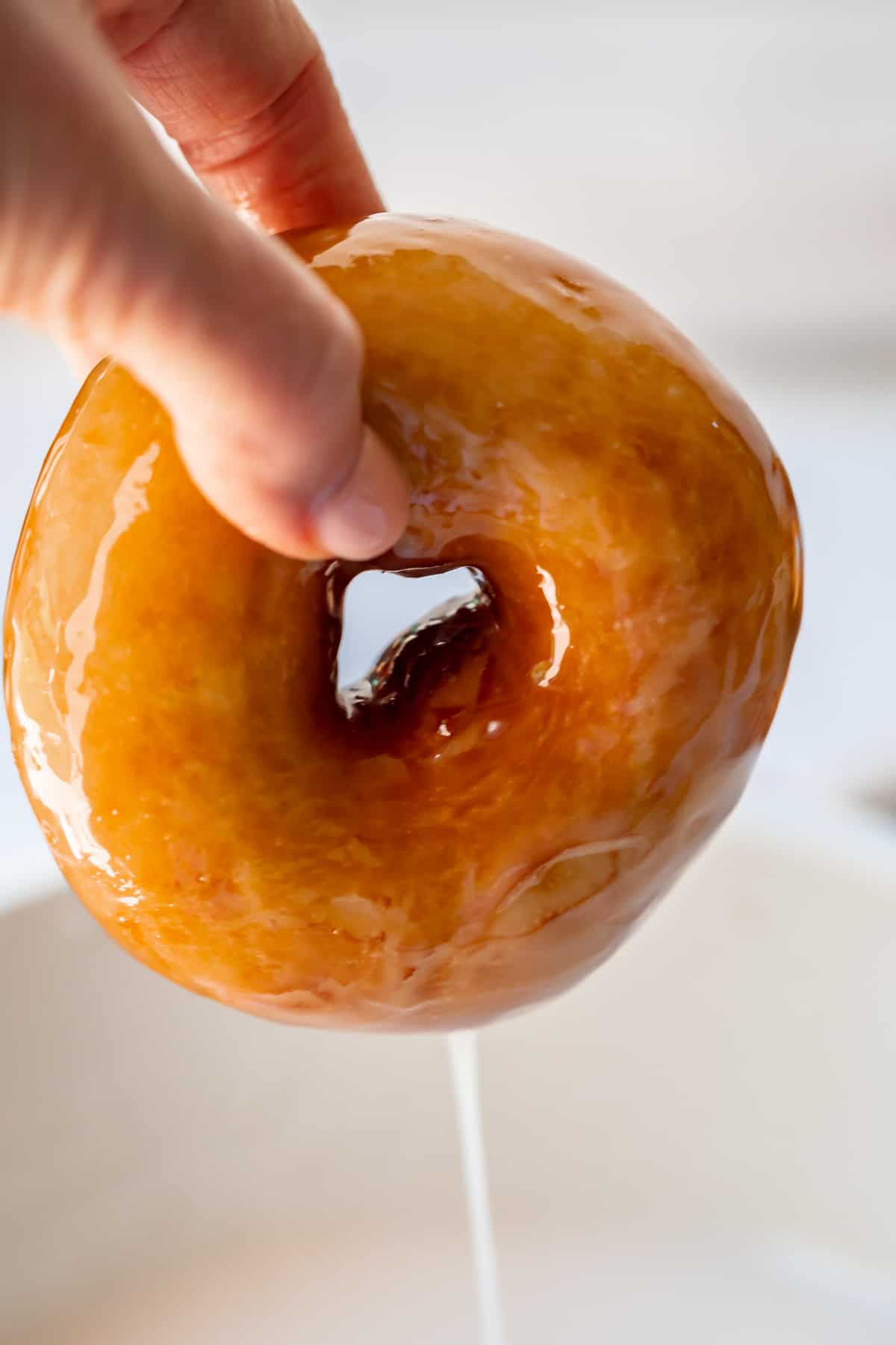 fingers pulling a donut fully coated in glaze out of the glaze bowl and letting the extra drip down.