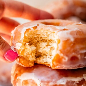 close up of hand reaching for a homemade glazed donut with a bite taken out of it.