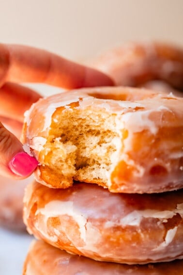 close up of hand reaching for a homemade glazed donut with a bite taken out of it.