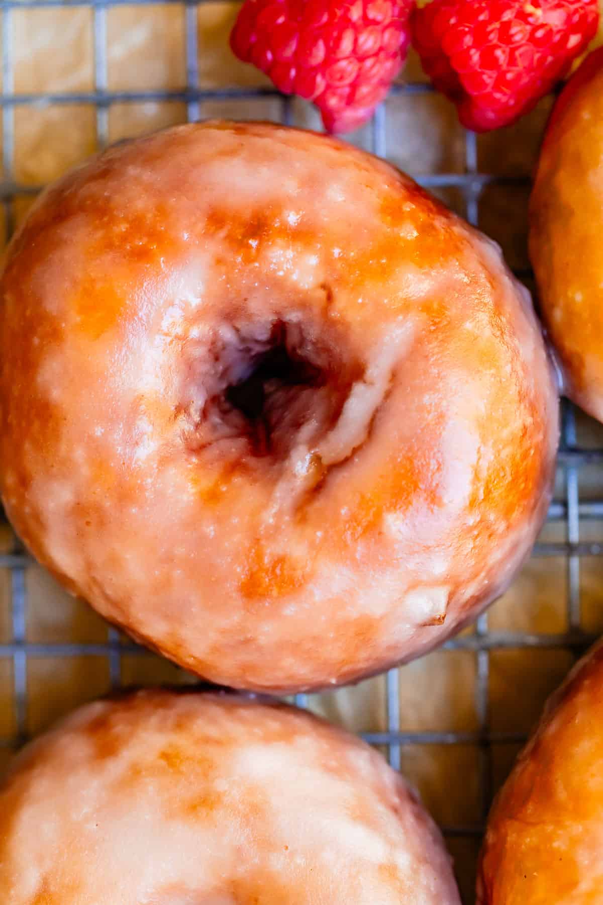 close up of one perfectly made from scratch glazed donut with raspberries next to it.
