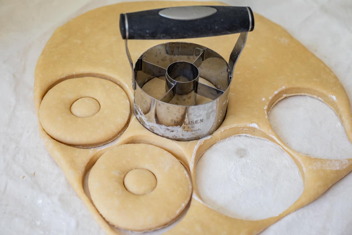 rolled out donut dough with a metal donut cutter cutting the donuts (and holes).