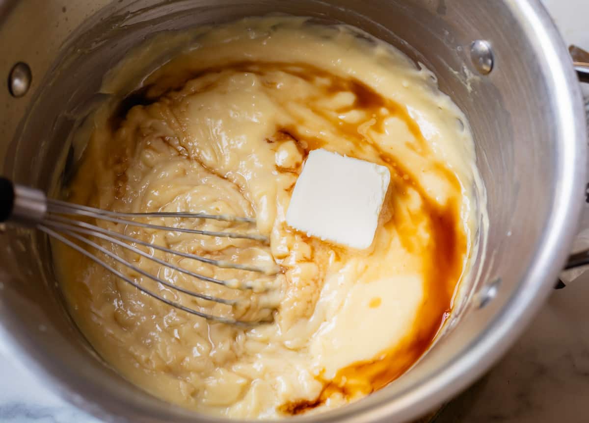 butter and vanilla being stirred into thickened pastry cream in a silver pot.