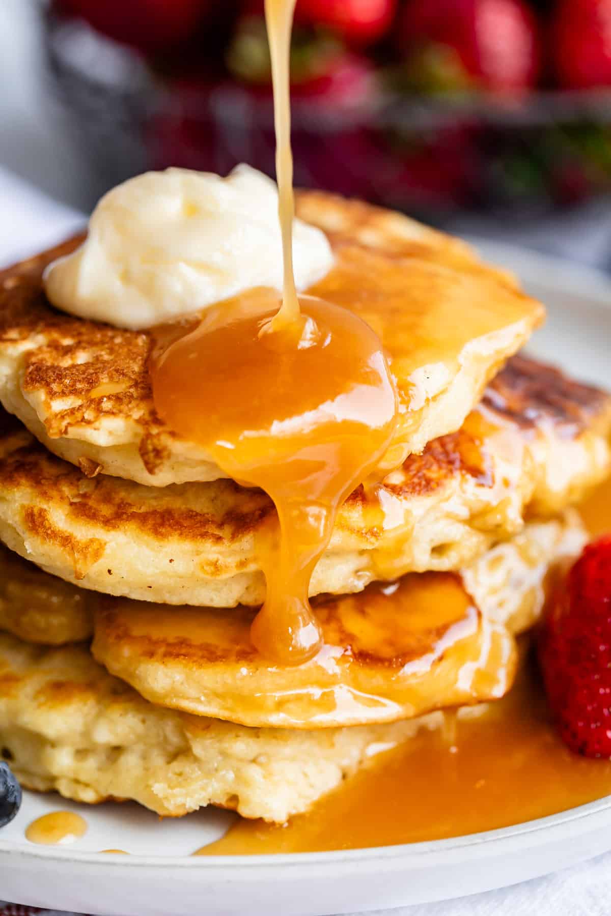 pouring buttermilk syrup over a stack of pancakes with butter and strawberry.