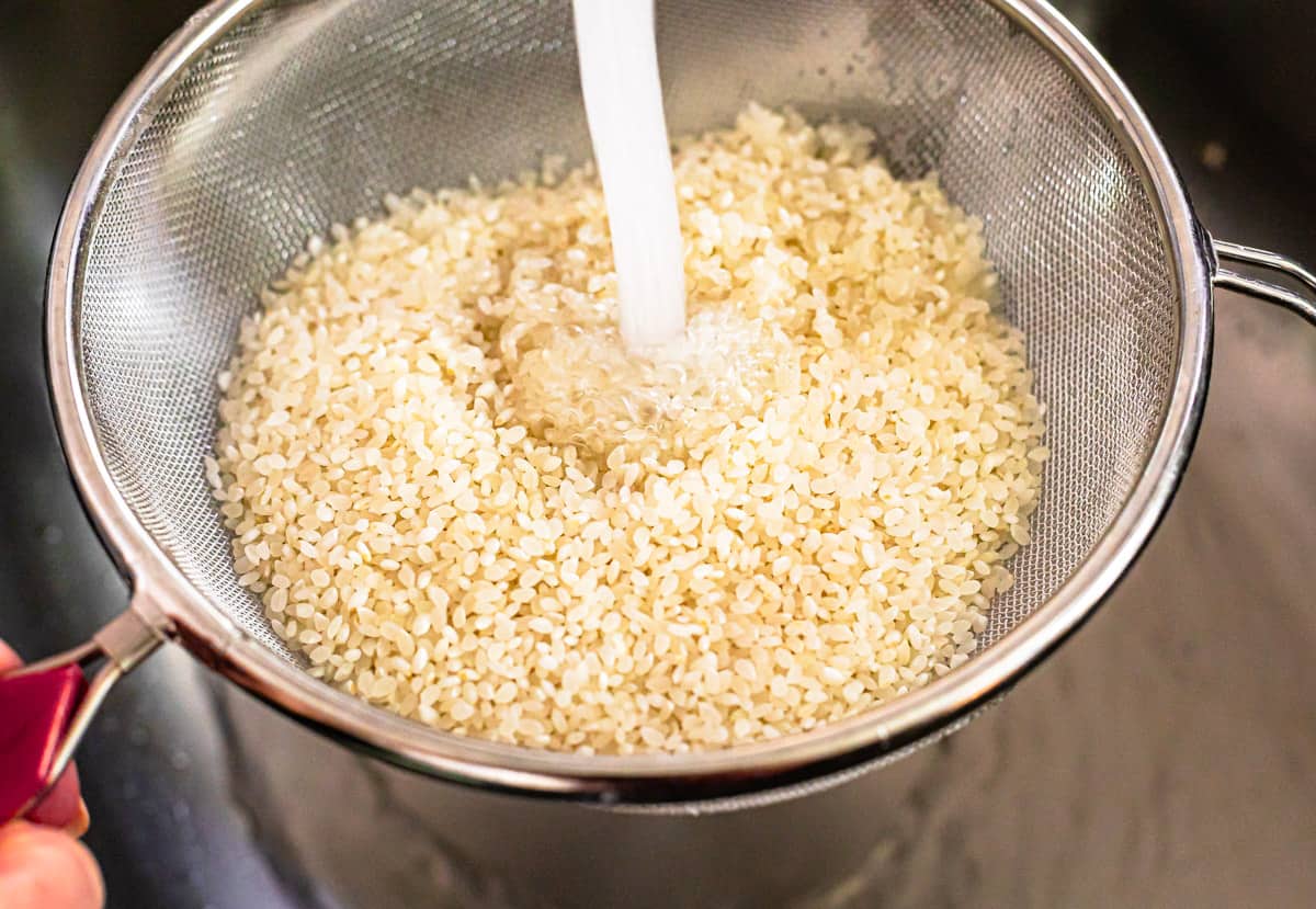 sushi rice in a strainer being rinsed with water from the kitchen faucet prior to cooking.