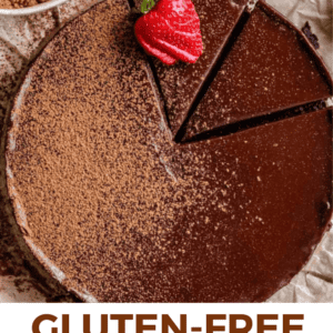 Round flourless chocolate cake topped with cocoa powder and strawberries, with two slices cut out. A bowl of cocoa powder and a single strawberry are nearby. Text reads Gluten-Free Chocolate Cake – The Food Charlatan.