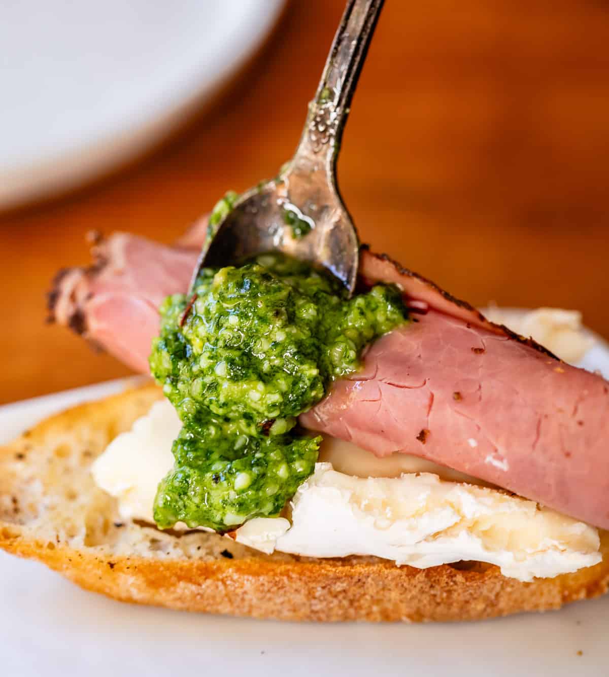 Arranging a bite of triple-cream cheese, deli-cut roast beef, homemade pesto, on a slice of baguette close up.