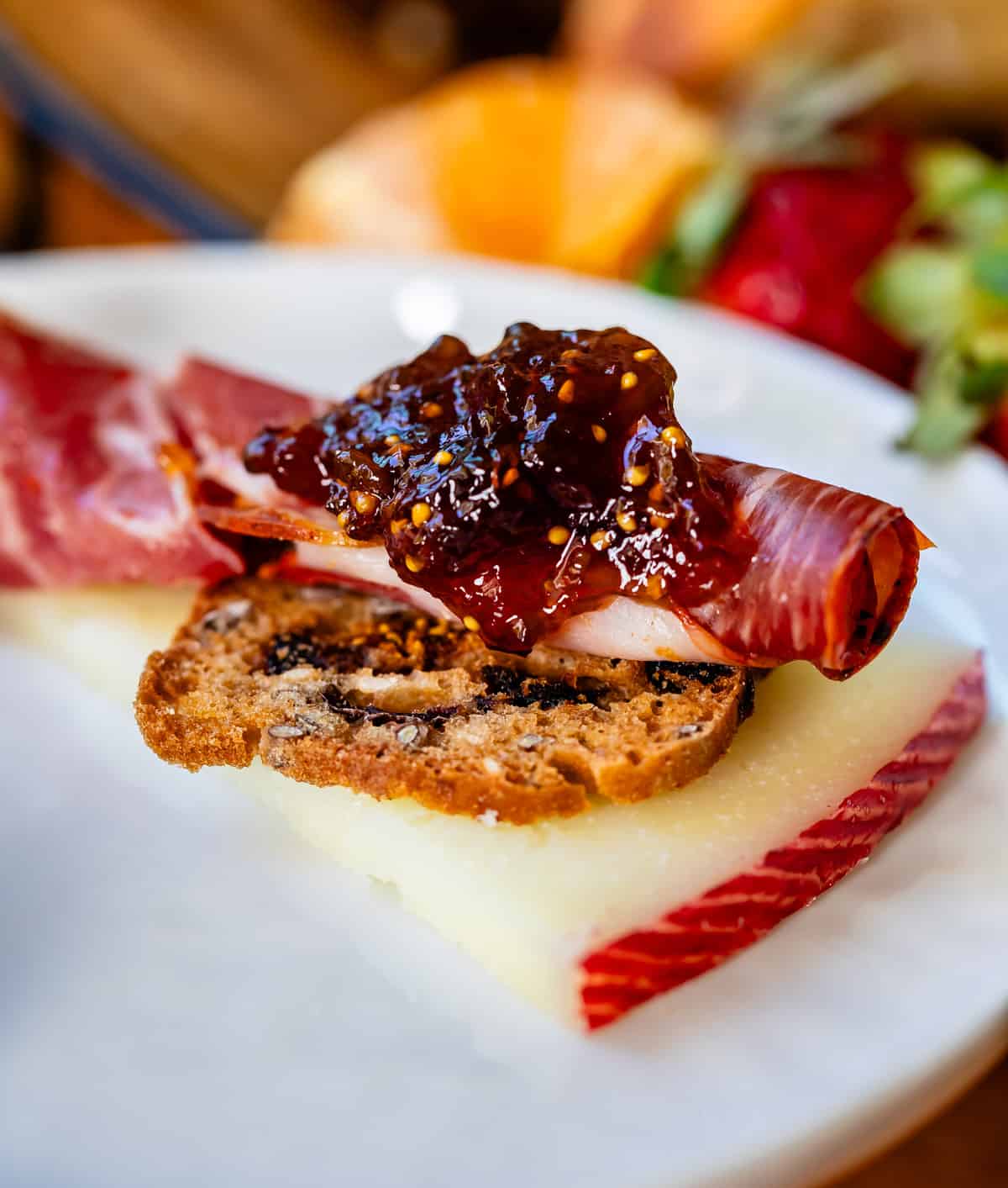 Delectable morsel of cheese, sweet cracker, deli meat, and fig jam close up on white plate.