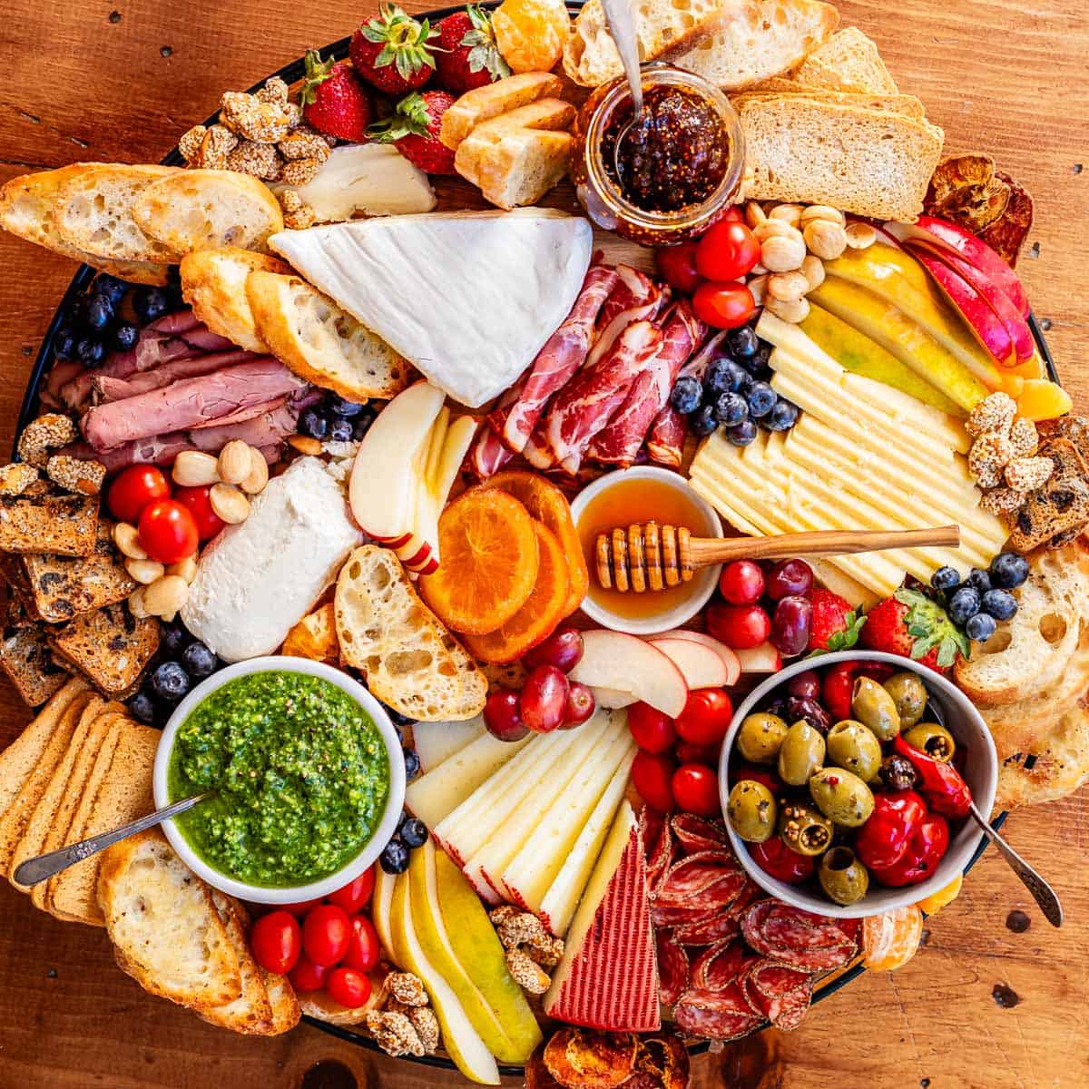 overhead shot of a round charcuterie board with brie, cheddar, oranges, pesto, crackers, bread, etc.