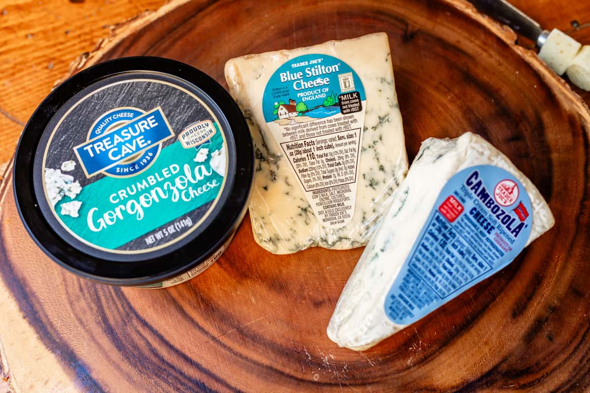 Arrangement of crumbly, stinky cheeses in packaging on wooden board including gorgonzola, blue stilton, and triple cream.