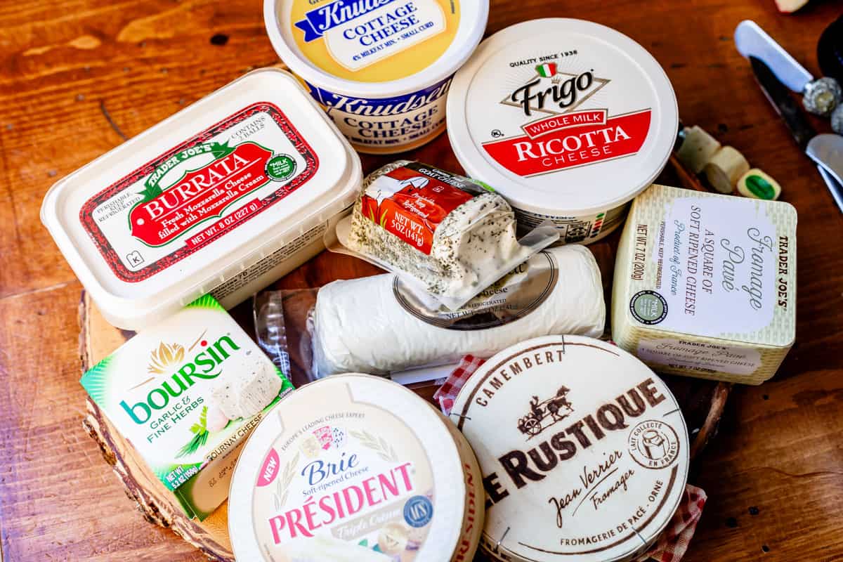 Arrange of soft cheeses in packaging on wooden board including burrata, boursin, brie, camembert, goat, ricotta, cottage, and pave.