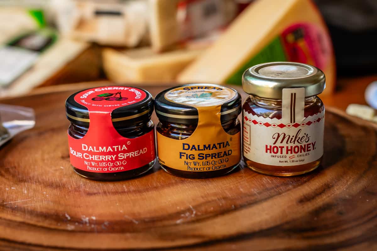 three small jars of spread including sour cherry, fig, and hot honey on wooden board for display.