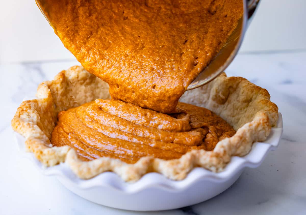 pouring sweet potato pie filling from a mixing bowl into a par-baked pie shell in a ceramic dish.