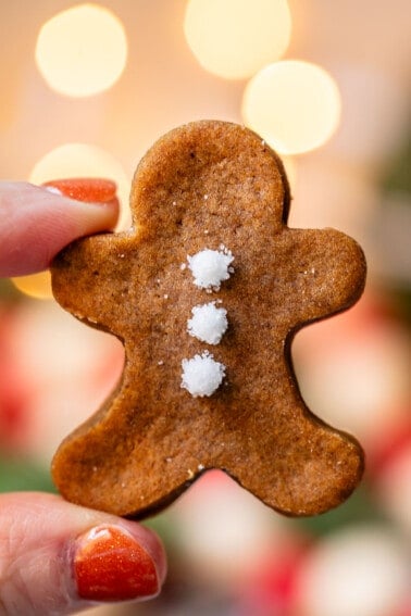 hand holding up tiny gingerbread man cookie with twinkle lights in background.