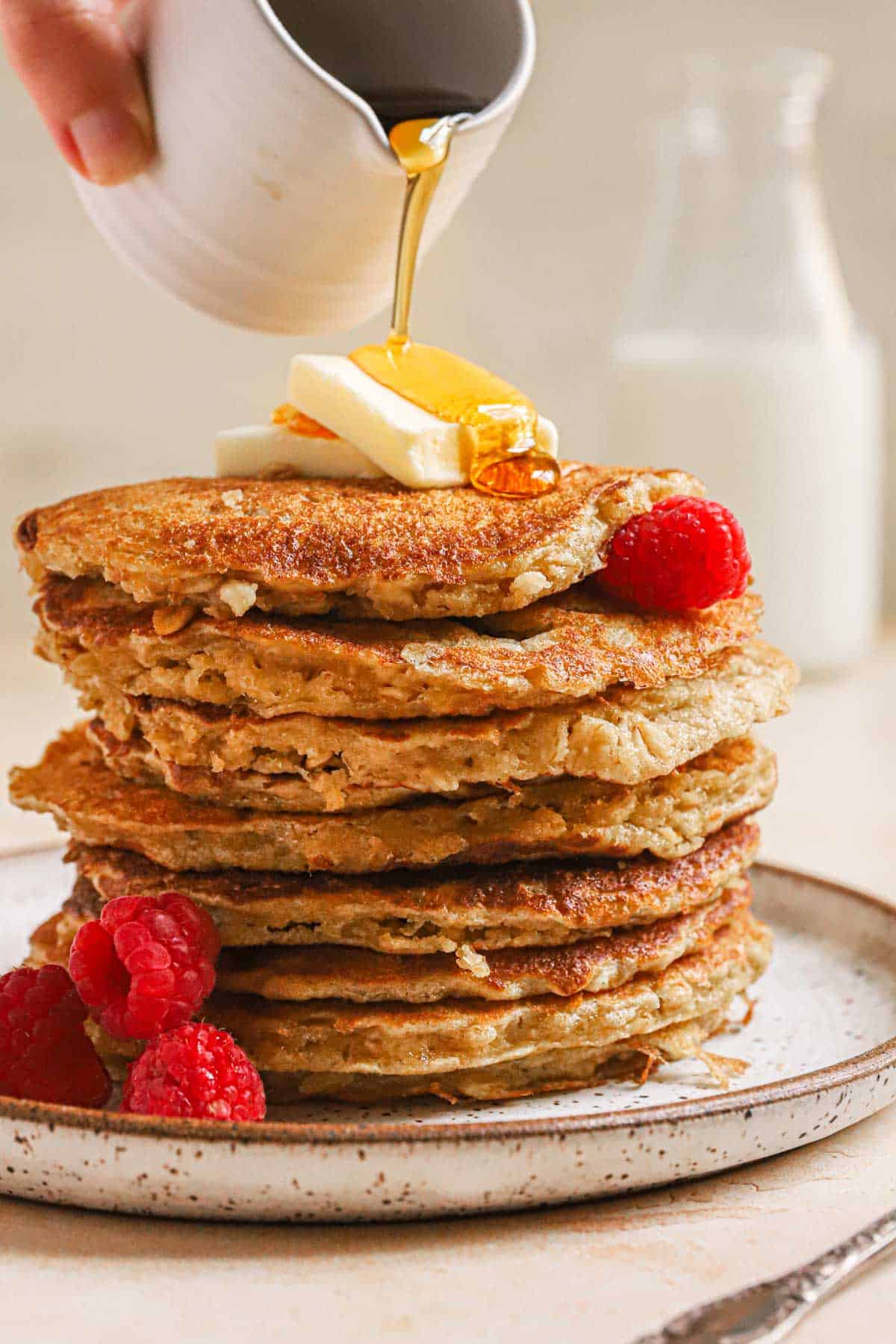 Large stack of oatmeal pancakes on ceramic plate with syrup being poured on top.
