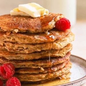Short stack of oatmeal buttermilk pancakes with raspberries and syrup drizzling down on ceramic plate.
