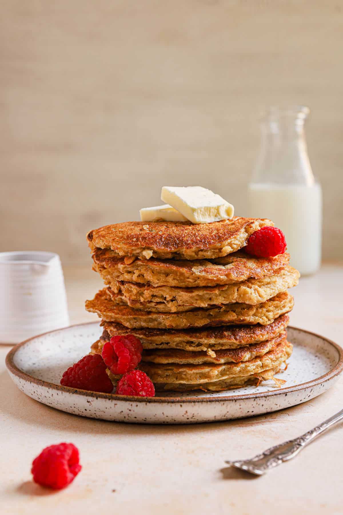 Stack of oatmeal pancakes on ceramic plate with raspberries and butter.