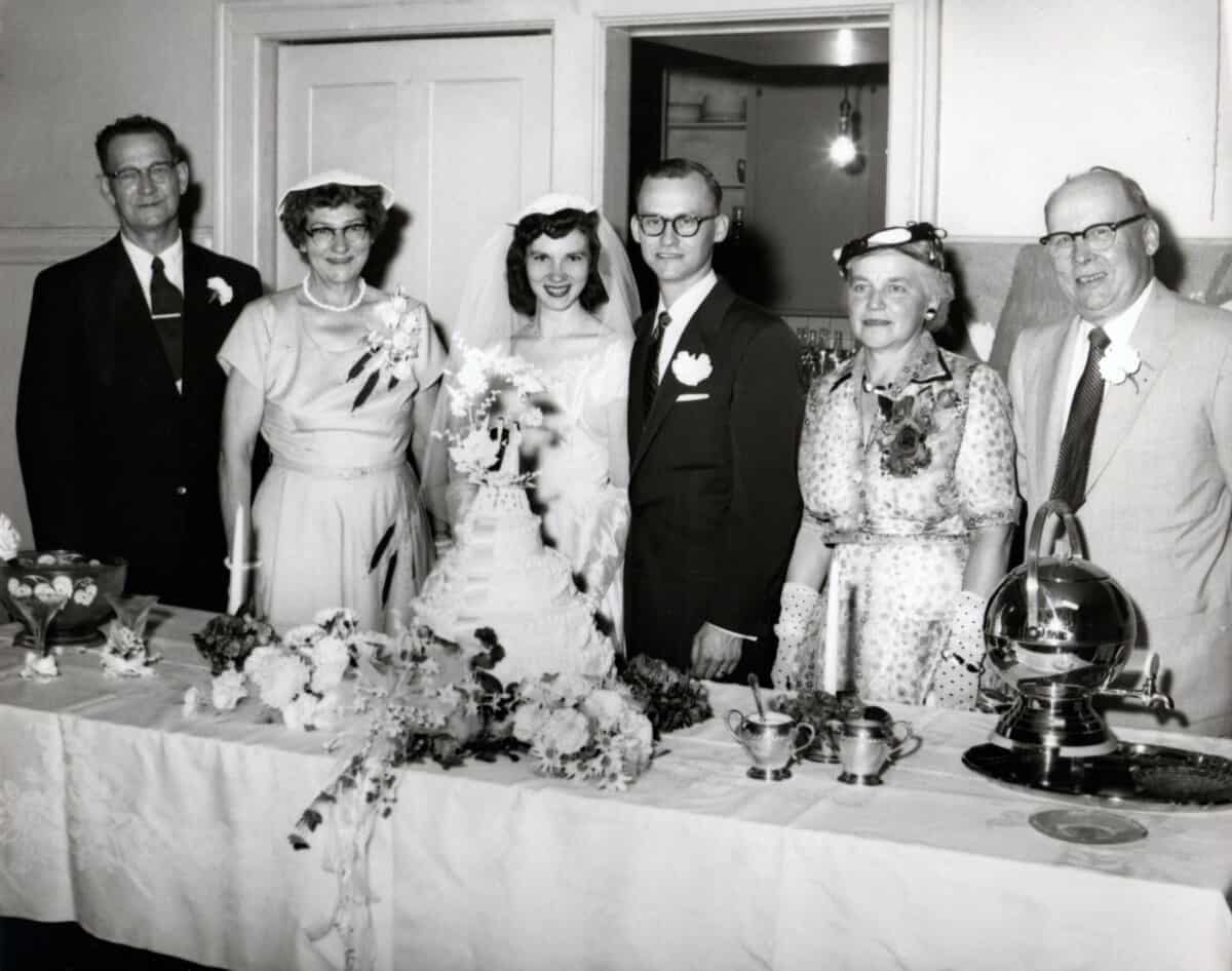 6 people standing behind a table with a wedding cake.