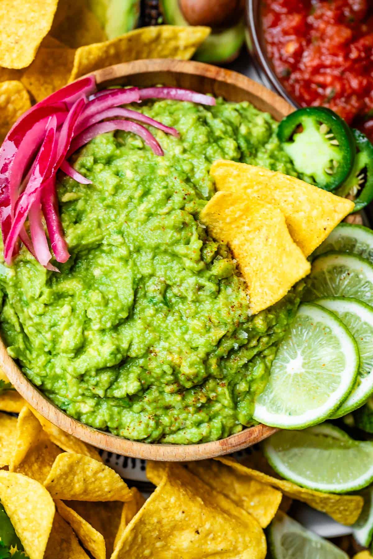 wooden bowl filled with guacamole and surrounded by chips, lime slices, and red onion.