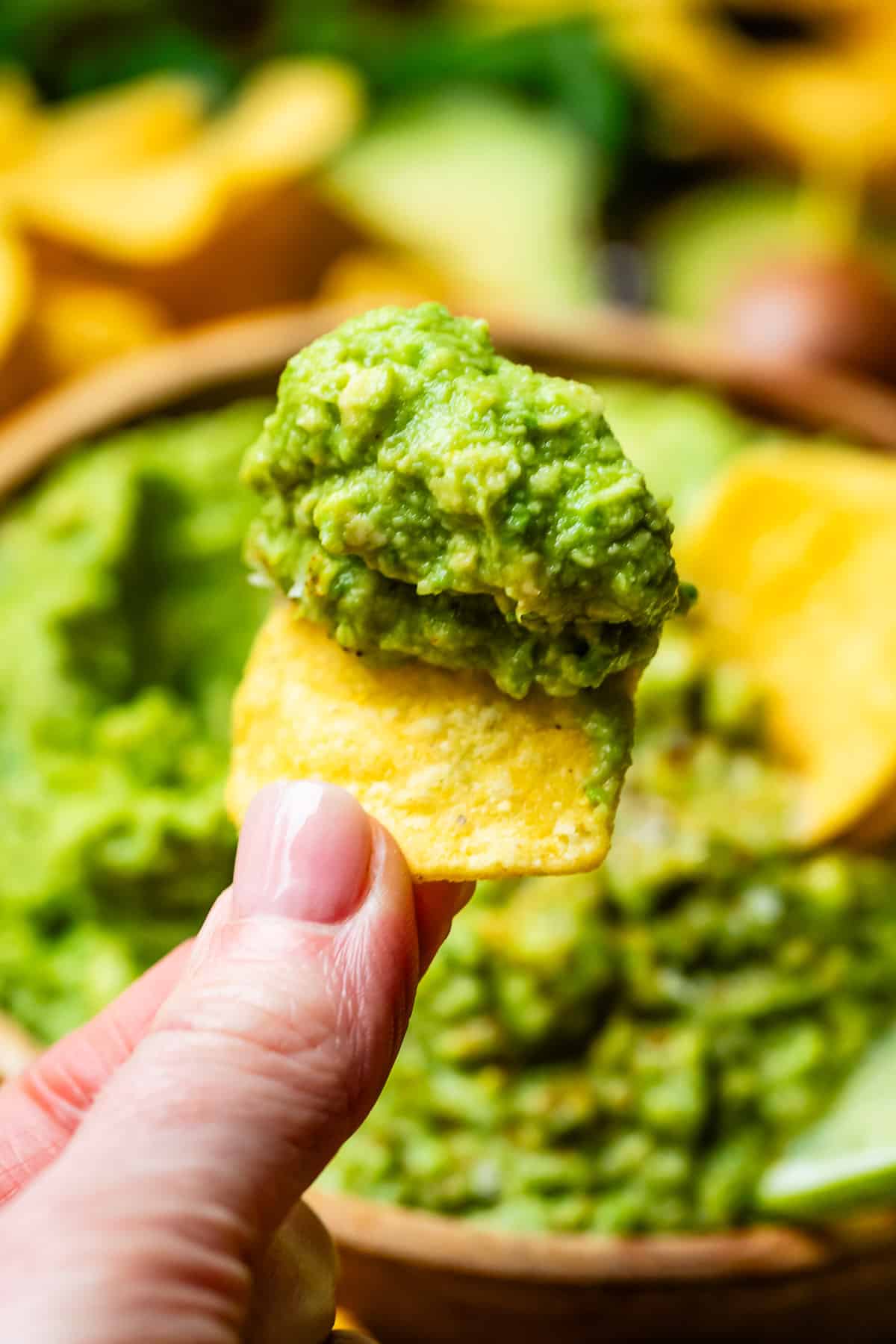 fingers holding a chip with a big heap of guacamole on it, with the guacamole bowl in the background.