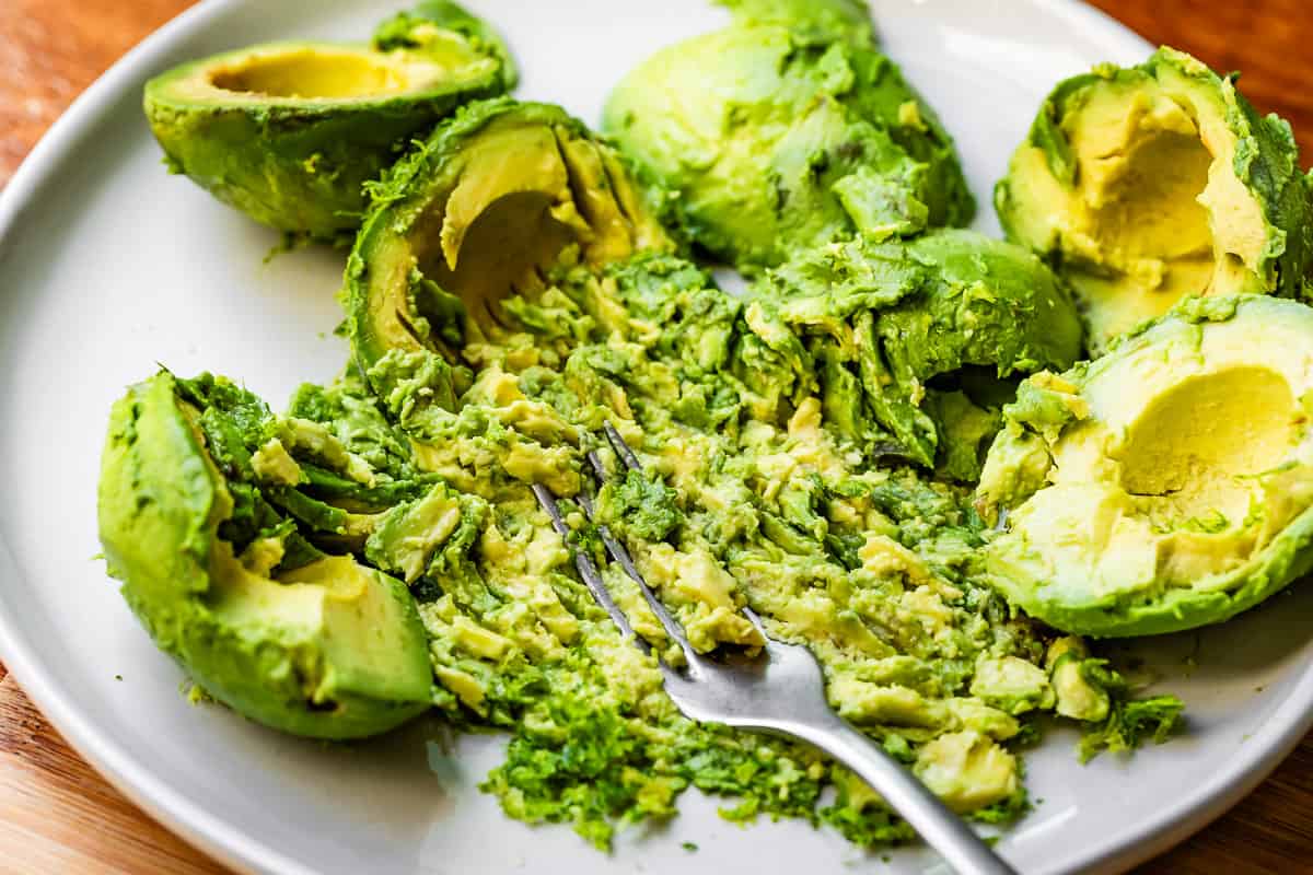 a metal fork mashing avocados on a plate; about one third of the avocado halves are smashed.