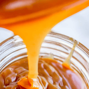 Close-up of caramel sauce being poured into a glass jar, with text above that reads, Homemade Caramel Sauce, Perfect for Desserts. The caramel looks smooth and glossy—the perfect finishing touch from your favorite caramel recipe.