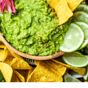 A bowl of homemade guacamole garnished with tortilla chips, lime slices, jalapeños, and pickled onions, surrounded by chips and a bowl of salsa. Text reads: Homemade Guacamole—your must-try fall dip recipe. The Food Charlatan.