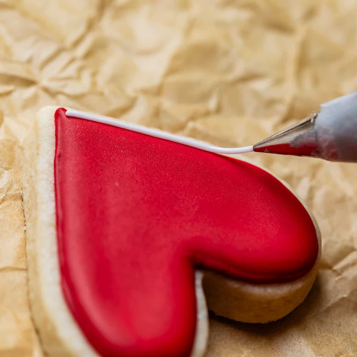 close up shot of a heart shaped cookie with red frosting on it, being lined with white piping.