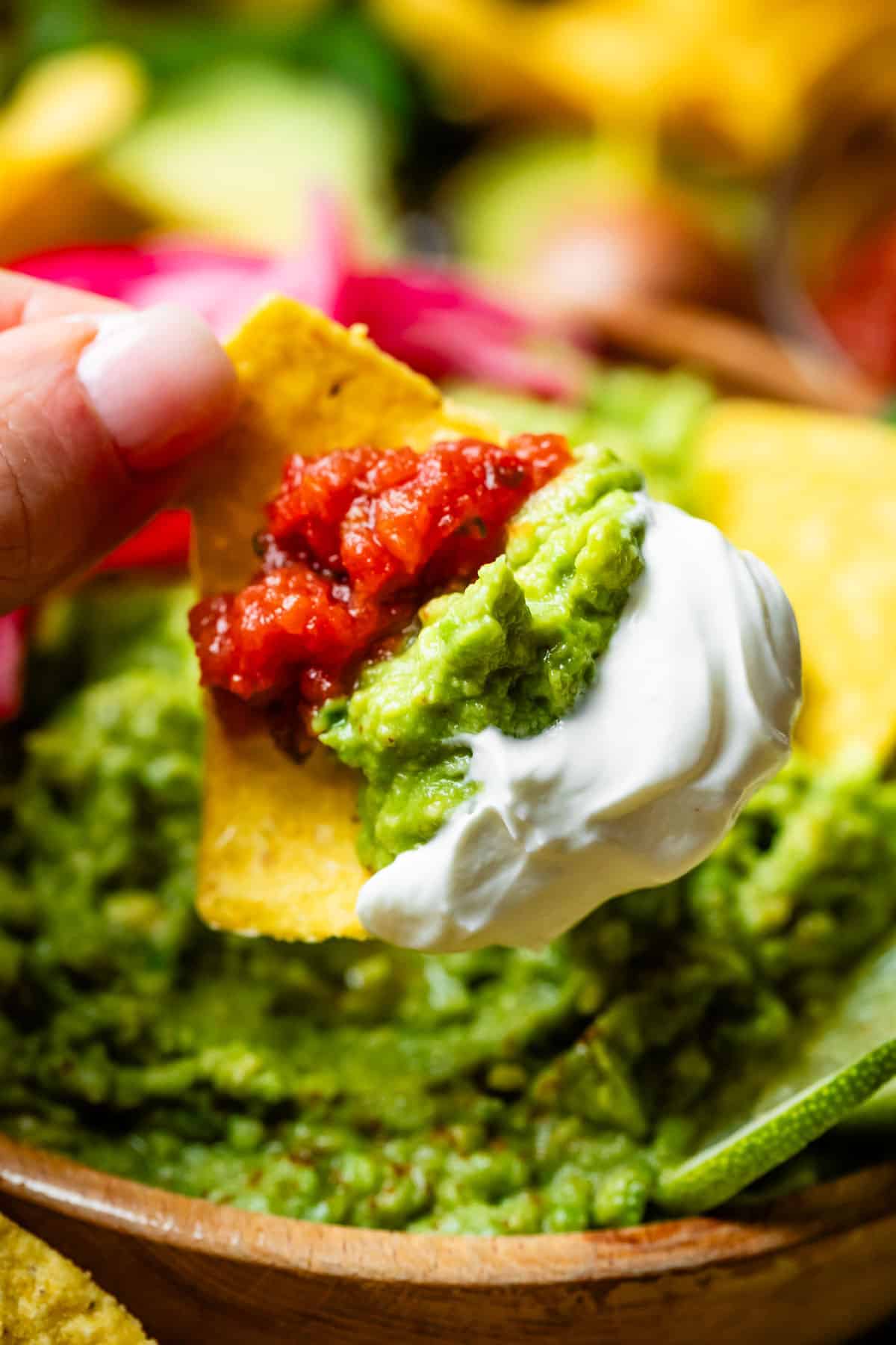 finger and thumb holding a chip loaded with guacamole, salsa, and sour cream.