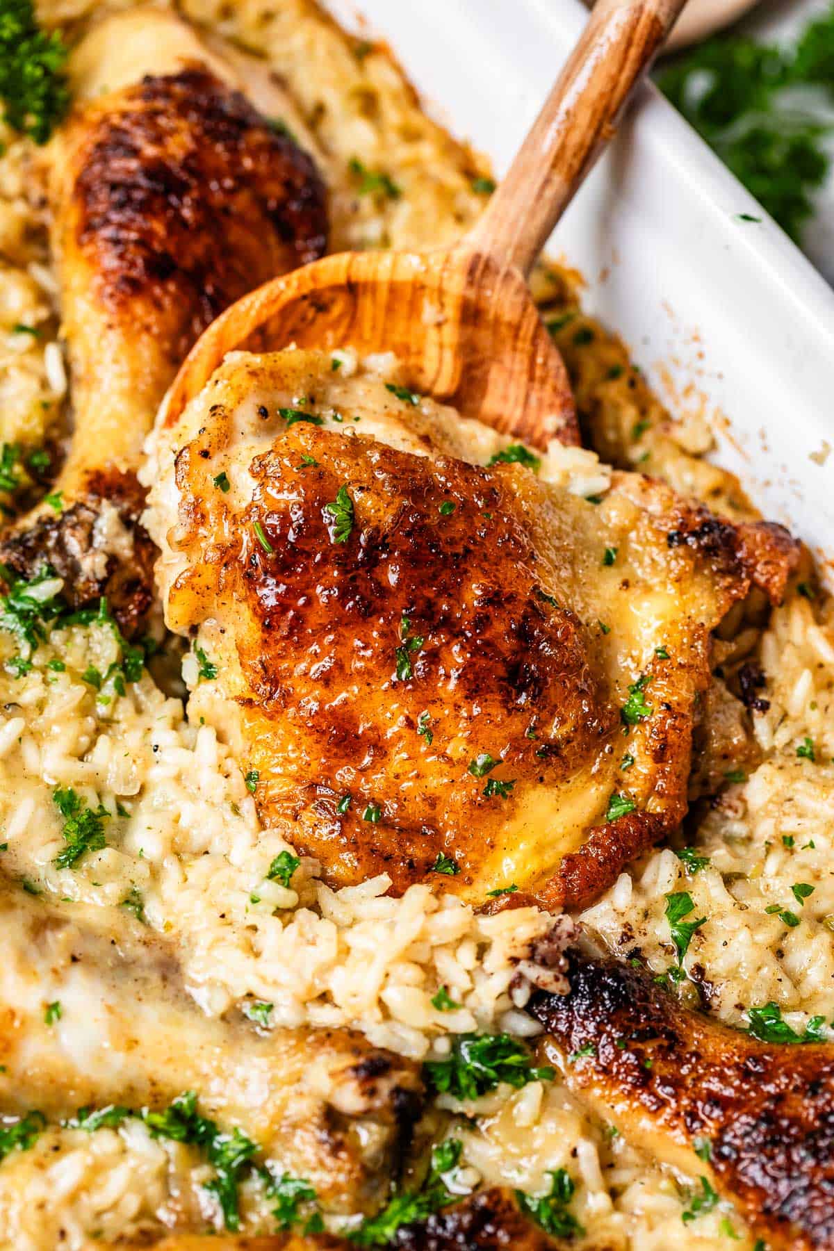 wooden spoon scooping up a cooked chicken thigh from the rice in a casserole dish.
