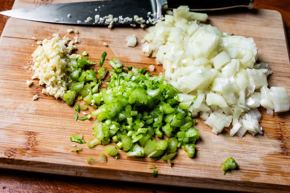 chopped onion, celery, and garlic on a cutting board with a chef's knife in the background.
