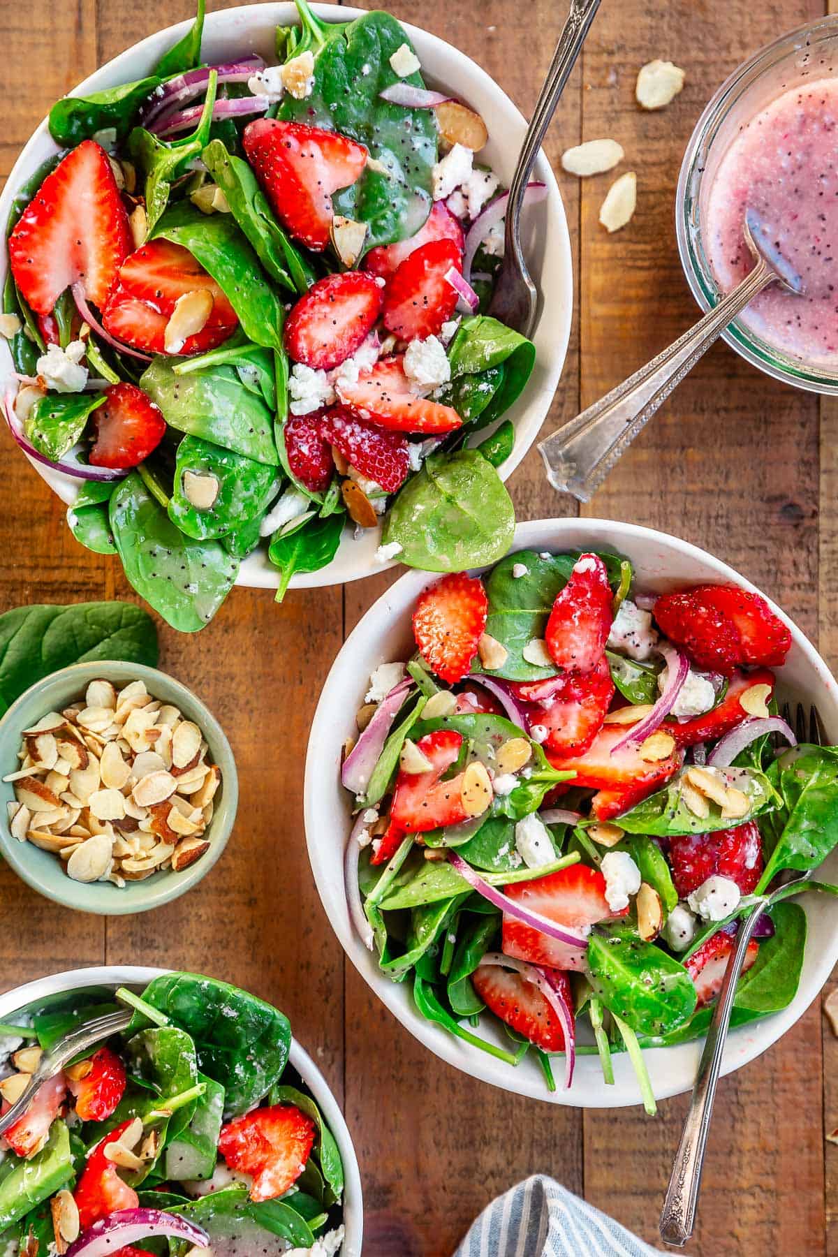 two bowls filled with dressed strawberry spinach salad with extra dressing and almonds on the side.