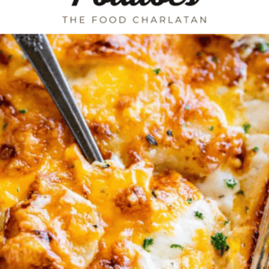 A close-up of scalloped potatoes in a baking dish, topped with melted cheese and garnished with herbs, with a serving spoon scooping a portion. Text above reads Cheesy Scalloped Potatoes - The Food Charlatan.