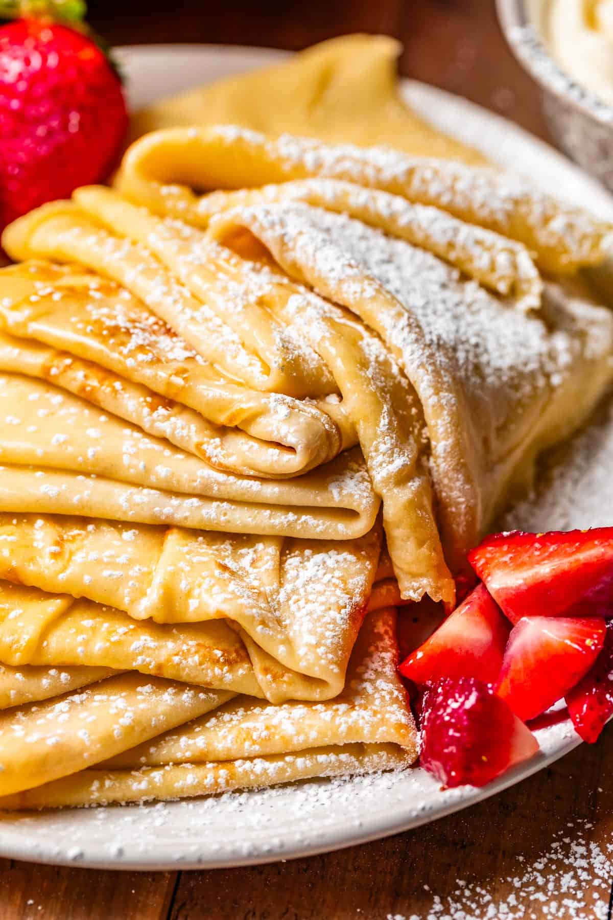 large stack of fresh folded crepes topped with powdered sugar and strawberries on the side.