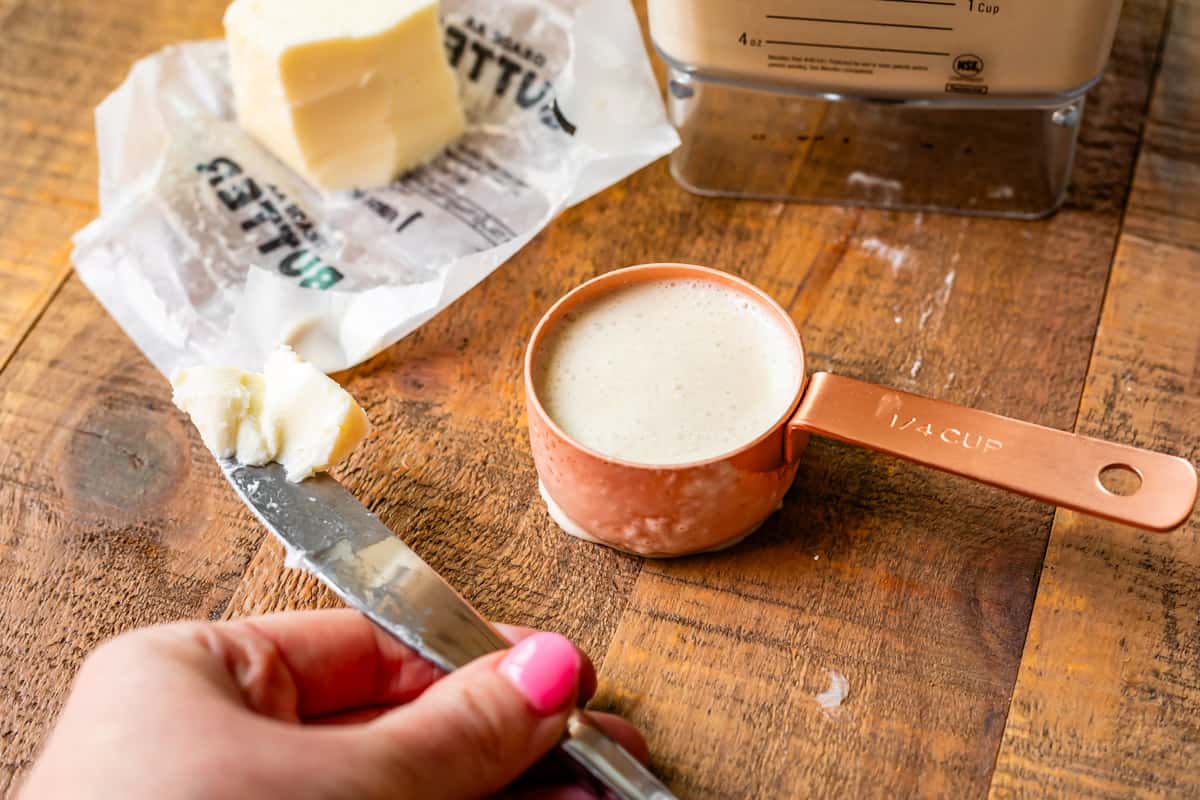 hand holding knife with butter on it and a measuring cup with batter in it on counter.
