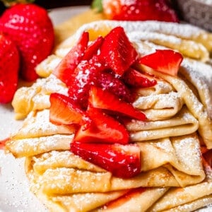 ceramic plate filled with several crepes topped with strawberry topping and powdered sugar.