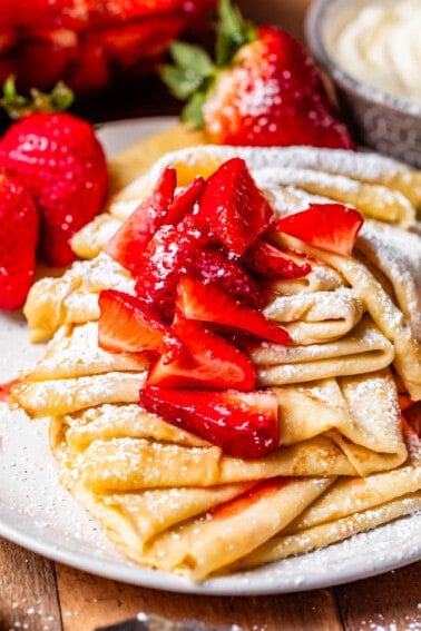 ceramic plate filled with several crepes topped with strawberry topping and powdered sugar.