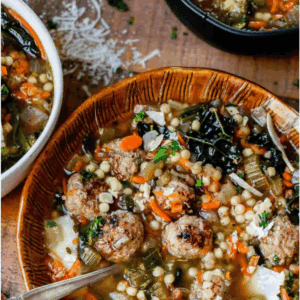 A bowl of hearty Italian wedding soup with meatballs, leafy greens, carrots, and pasta sits beside another bowl and grated cheese on a rustic wooden table, garnished with a red striped napkin and a spoon.