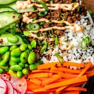 bowl brimming over with sections of rice, Korean beef with Sriracha mayo, veggies, and sesame seeds.