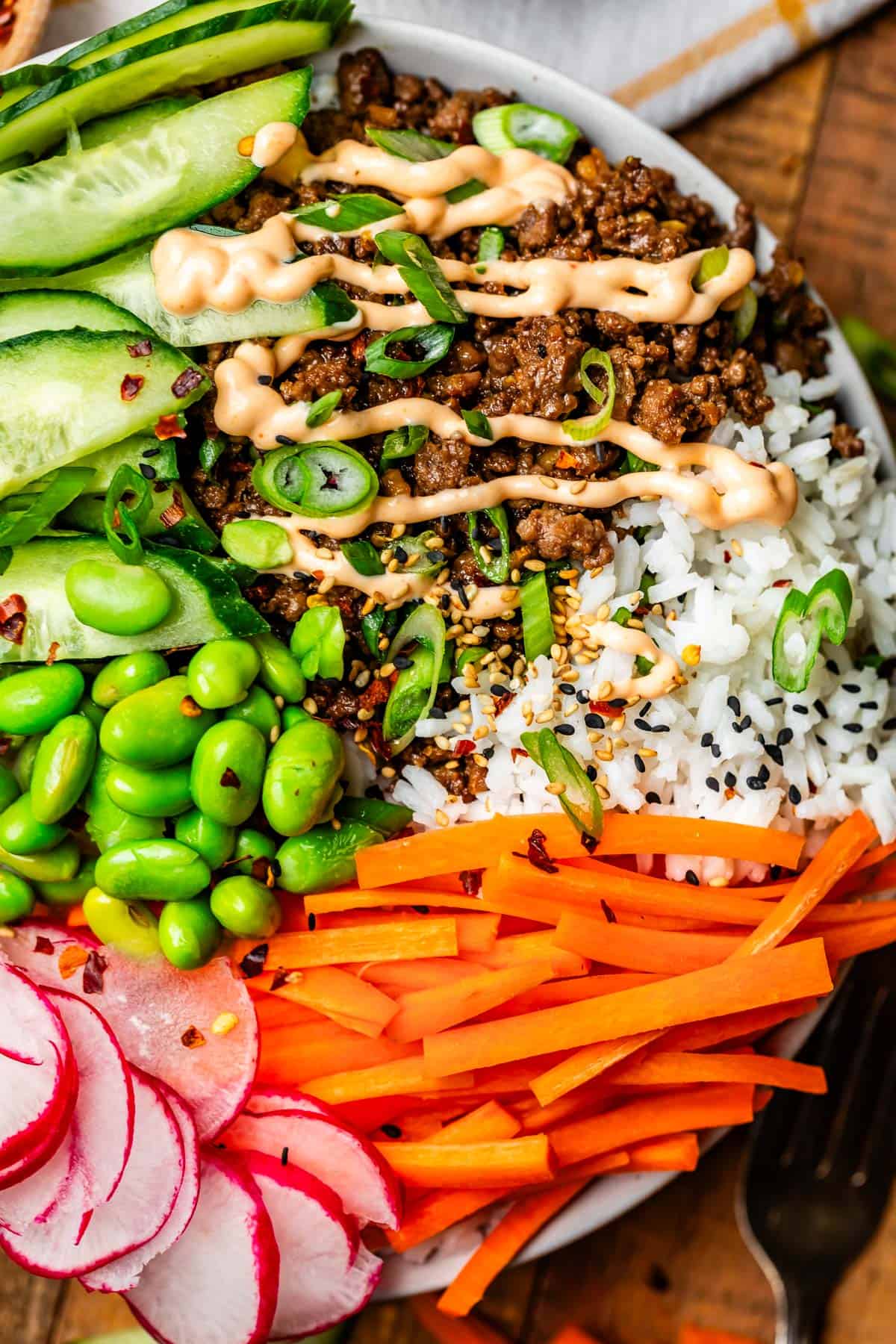 bowl brimming over with sections of rice, Korean beef with Sriracha mayo, veggies, and sesame seeds.