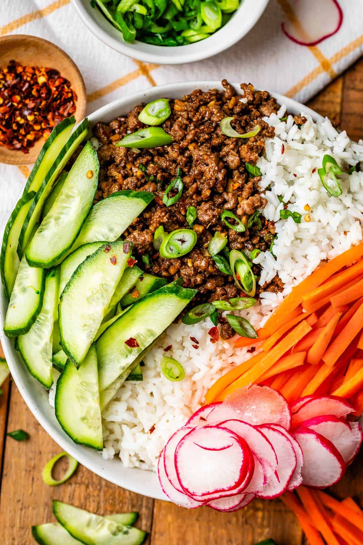 Korean beef bowl with rice and veggies on a wooden table with bowl of red pepper flakes.