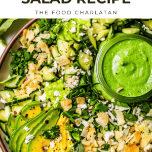 A vibrant green goddess salad topped with sliced avocado, crumbled cheese, crunchy toppings, and a small bowl of green dressing. Text above reads Green Goddess Salad - The Food Charlatan.