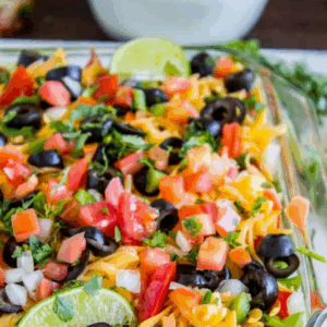 A close-up of a glass dish filled with colorful 7 layer dip, topped with shredded cheese, chopped tomatoes, onions, olives, cilantro, and lime slices. Text above reads, 7 Layer Bean Dip.