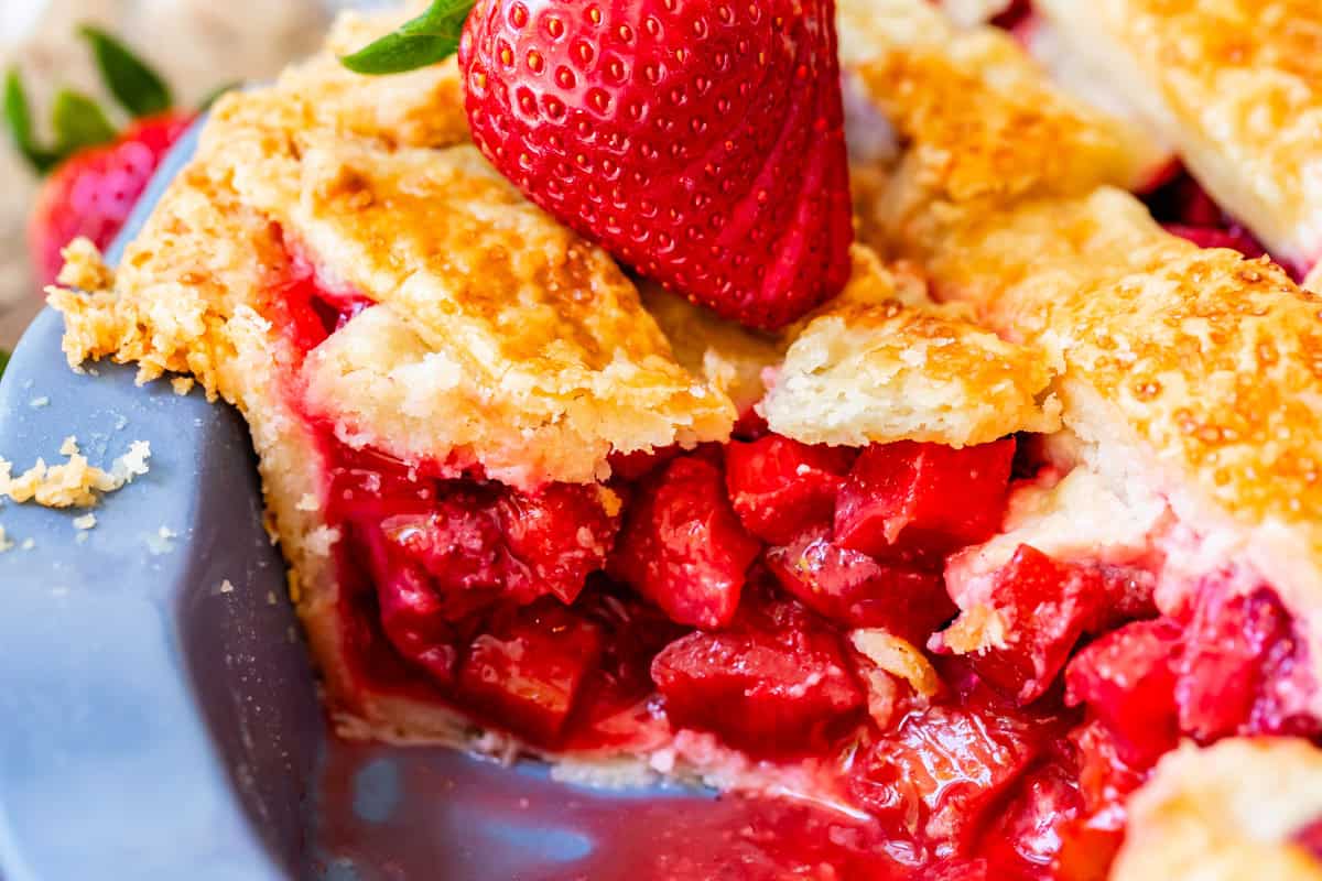 close up of side view of a slice of strawberry rhubarb pie with flaky crust and strawberry garnish.
